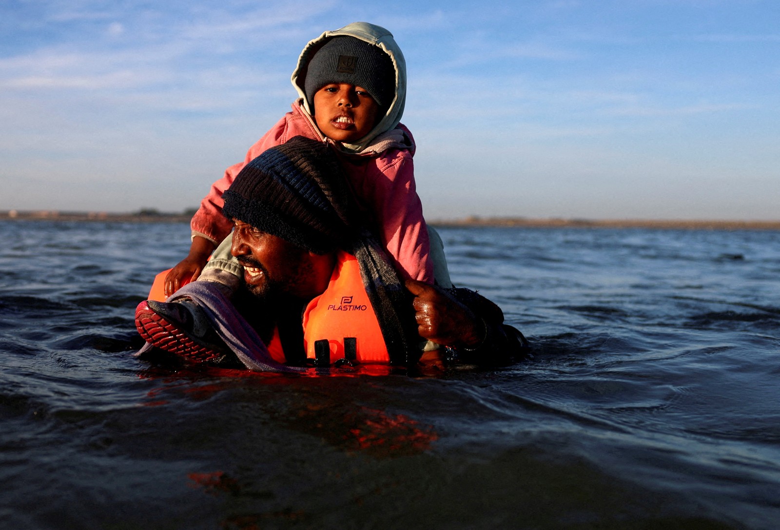 A person wearing a lifejacket, standing in chest-deep water, carries a child on their shoulders.
