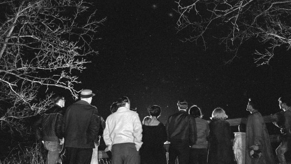A black-and-white photograph of a group of people looking up at the night sky, which is framed by some trees