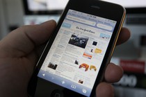 A left hand holds an iPhone with The New York Times' homepage displayed on the screen.