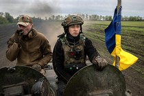 Two members of the Ukrainian 28th Mechanized Brigade ride in an armored vehicle towards hostile Russian forces. The one on the left is smoking a cigarette, the one on the right is scowling, and to their side is a blue and yellow flag.