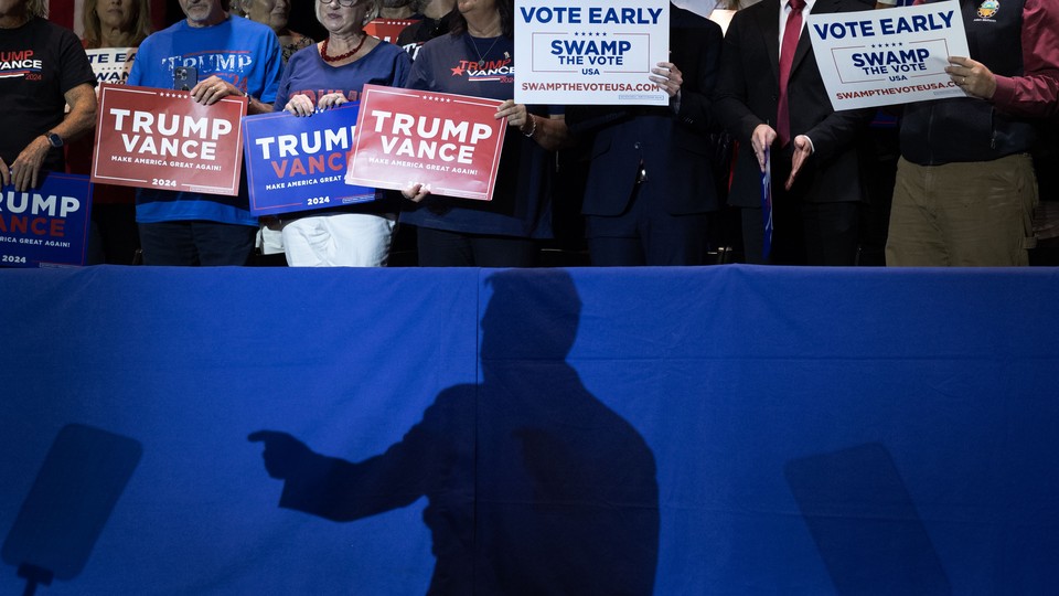 The dark shadow of a man is projected onto a blue barrier. Behind it, attendees at a Trump rally hold signs.