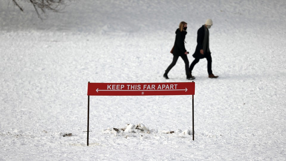 Two people walk together in a snowy NYC park behind a sign that says: "Keep this far apart."
