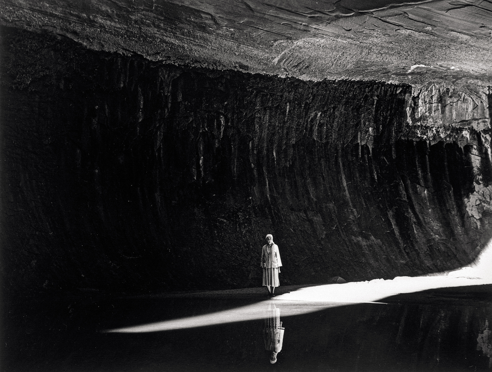 black-and-white photo of O'Keeffe standing in middle of photo in a horizontal beam of sunlight with canyon above and her mirrored reflection in the water below