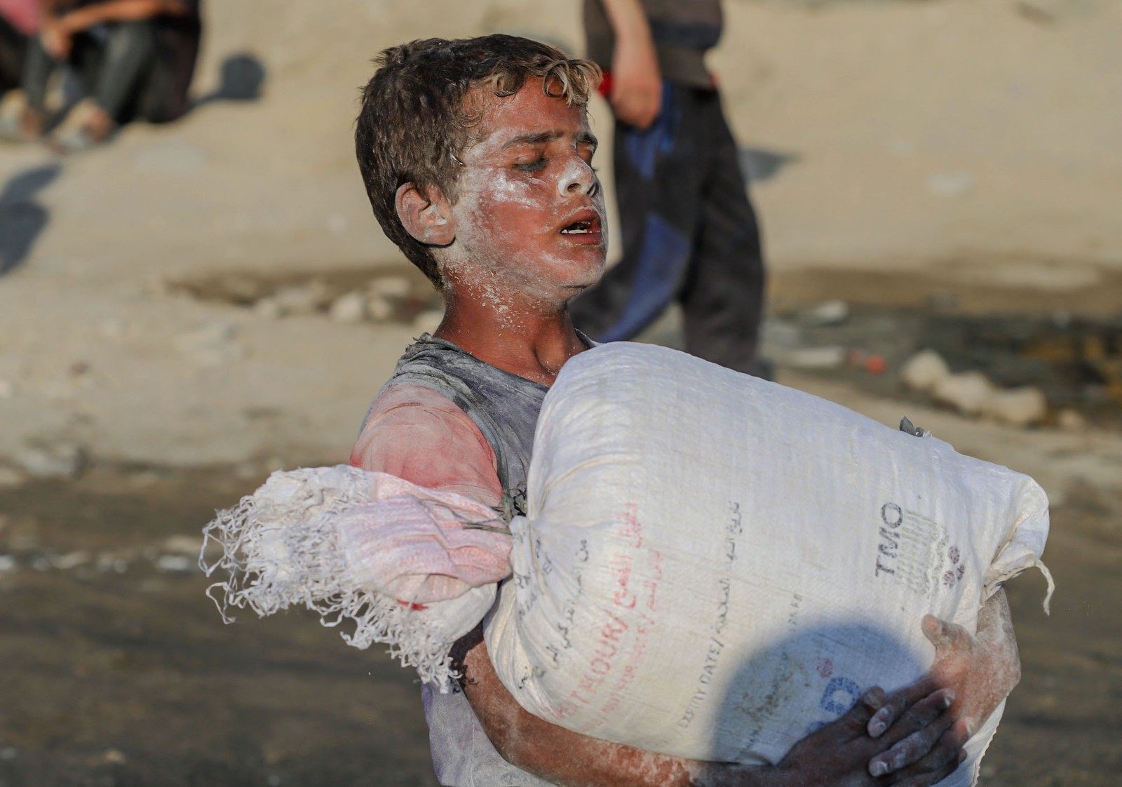 A boy with flour smeared on his face struggles to carry a large bag of flour.
