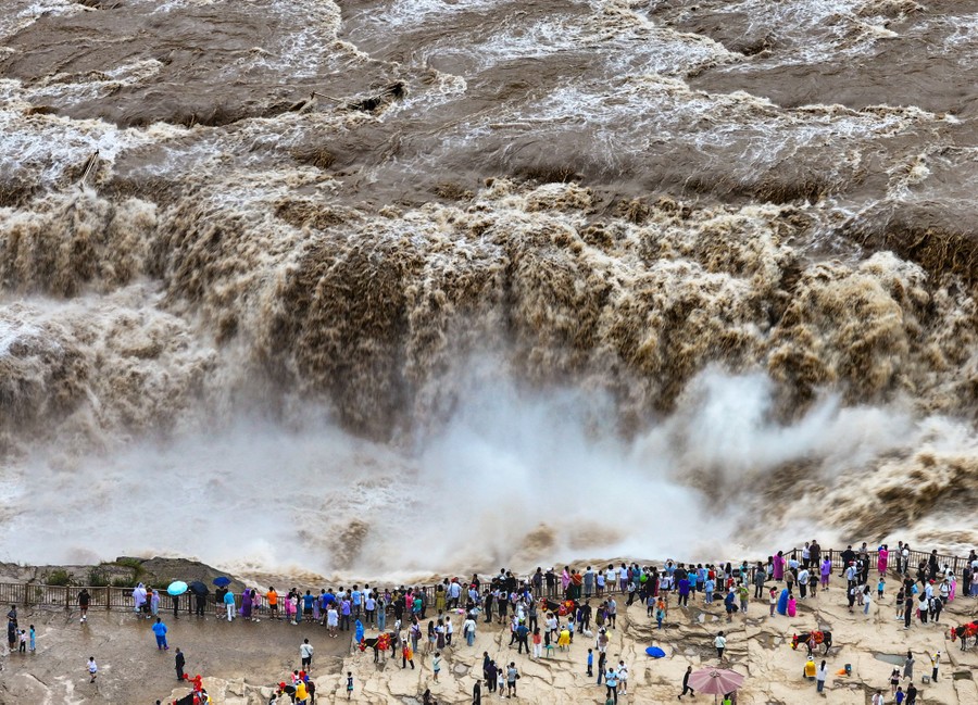 Tourists stand in a viewing area, looking out over a rushing waterfall.