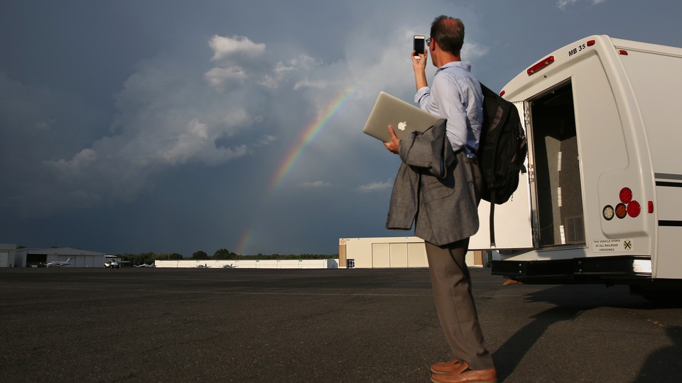 A man in business clothes carrying a laptop and a backpack uses a smartphone to take a picture of a rainbow.