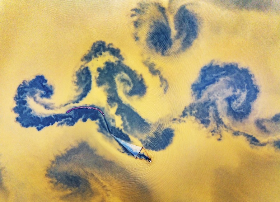 An aerial view of a fisherman in a small boat on yellow-colored lake water, leaving dark spiral swirls in its wake