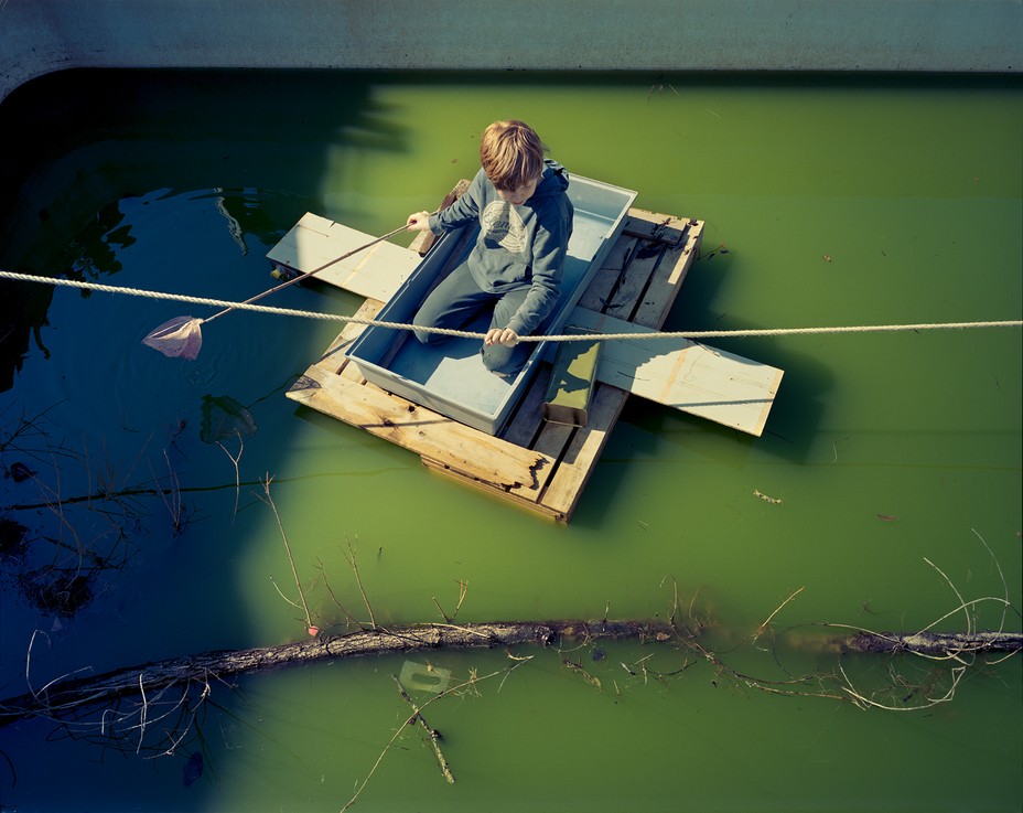 boy holding a rope and net in homemade boat on murky green water 