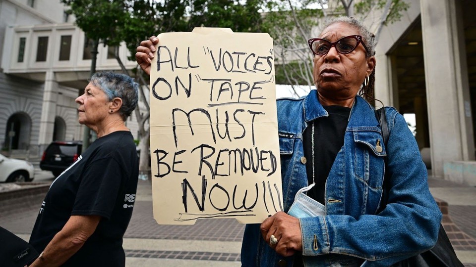 A woman holding a sign that reads "All 'voices' on 'tape' must be removed now!!!"