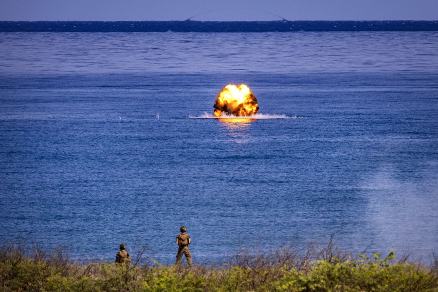 Two soldiers stand on shore, looking out toward an explosion on the ocean's surface.