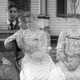 One man and two women in clothes from the past, all semi-transparent like ghosts in front of a house's porch