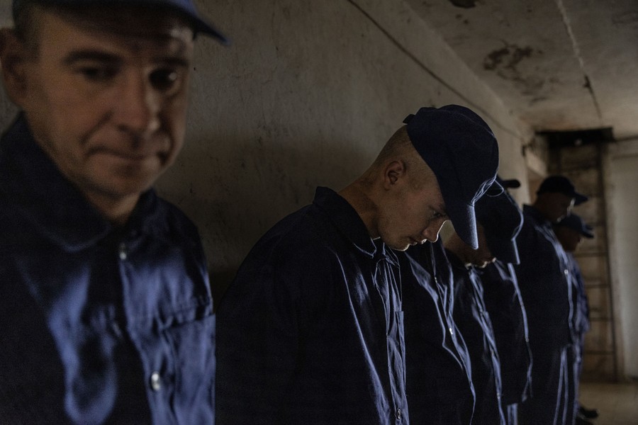 A half dozen men in blue prisoner uniforms stand against a concrete wall.
