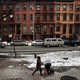 A woman pushes a stroller down a sidewalk lined with brownstones.