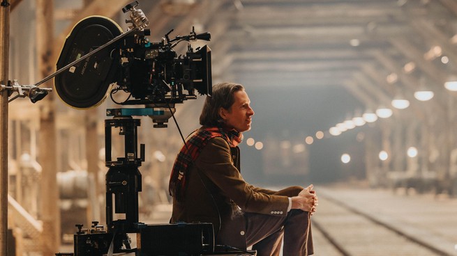 A behind the scenes image of Wes Anderson filming, wearing a brown suit and a red plaid scarf.