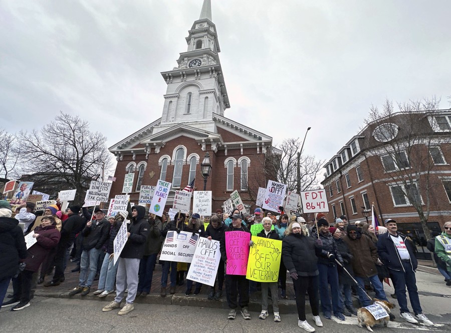 A group of protesters holding signs stands outside of a historic building in New Hampshire.