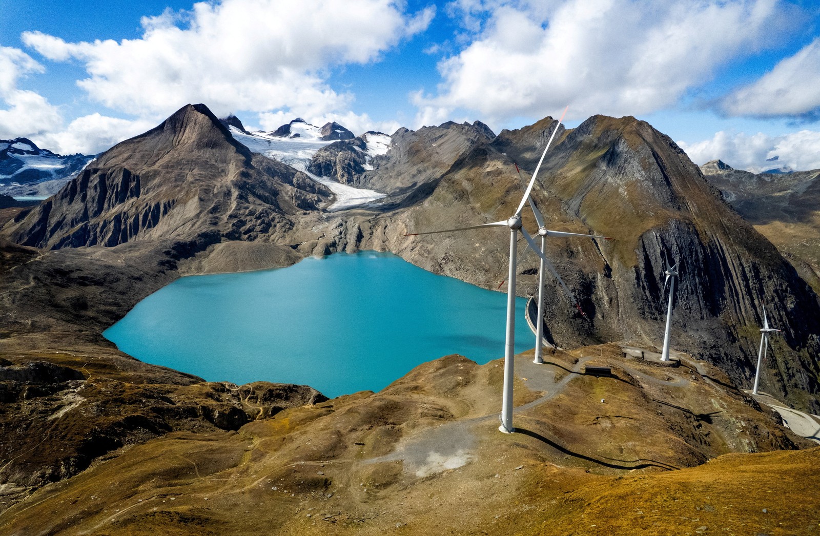 A reservoir in a steep mountain landscape, with a glacier and several wind turbines visible.