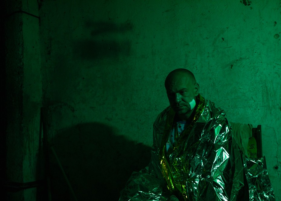 A Ukrainian solder sitting in a dark room  with green lighting, wrapped in a thermal blanket with a bandage on his face.