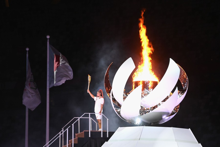An athlete stands before a fire in a large ornamental cauldron, holding a ceremonial torch.