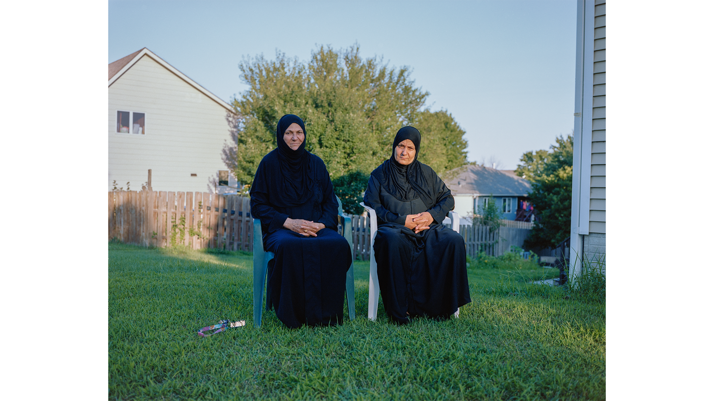 two women in hijabs and long-sleeved black dresses sit, hands in laps, in lawn chairs on green grass in yard next to house