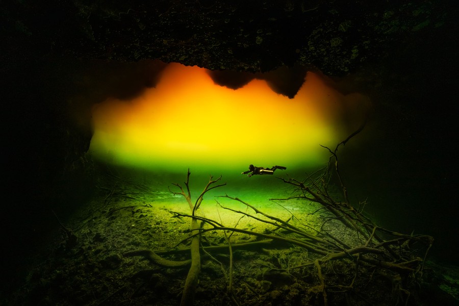 A diver swims past jagged rock formations, through water that varies in color above them, from green to yellow to red.