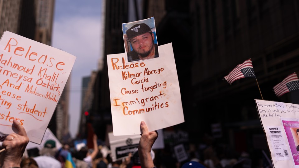 A hand holds up a sign that reads “Release Kilmar Abrego Garcia / Cease targeting Immigrant Communities” at a protest where others wave signs and upside-down American flags
