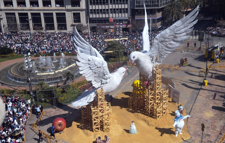 A crowd gathers in a city square to look at a large sculpture of two doves.