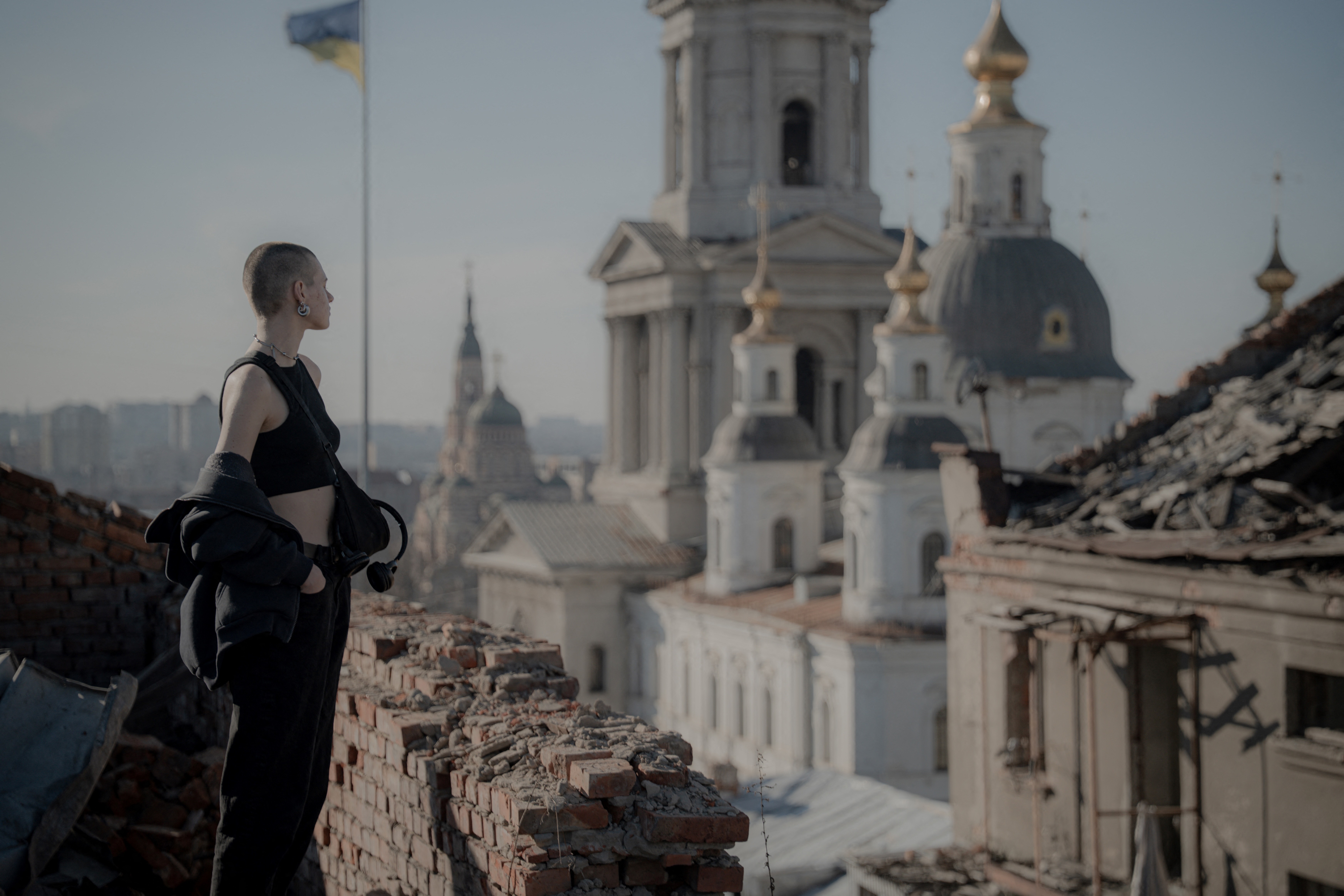 A civilian stands on a damaged rooftop overlooking a church and other large damaged buildings.