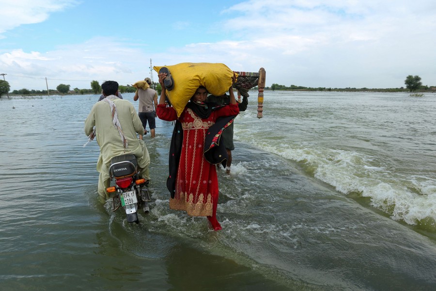 People carry belongings through shallow flowing floodwater.