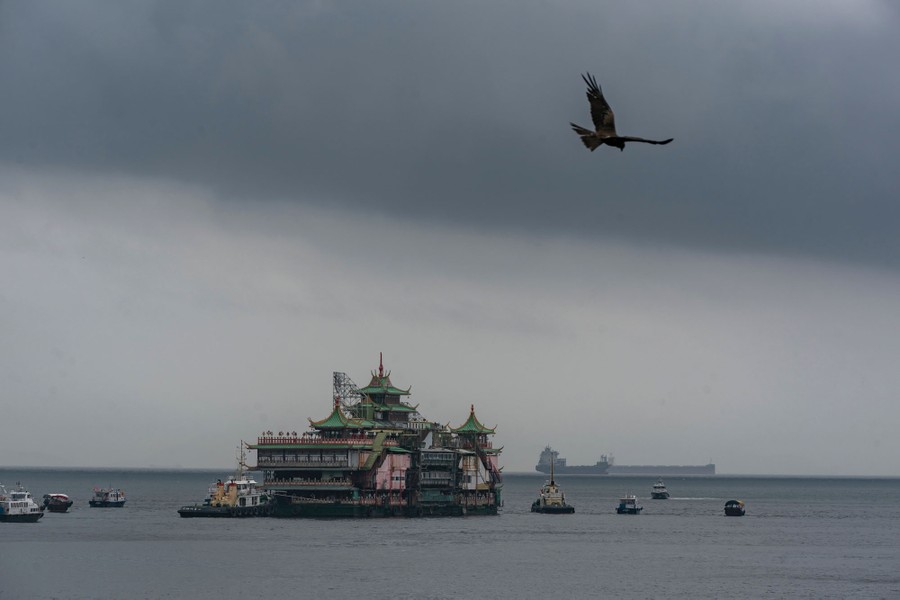 An ornate, several-story-tall floating restaurant is moved by tugboats.