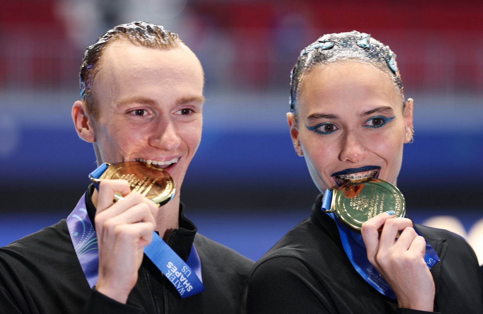 A pair of artistic swimmers smile while posing for the camera and biting their medals at a gold medal ceremony.