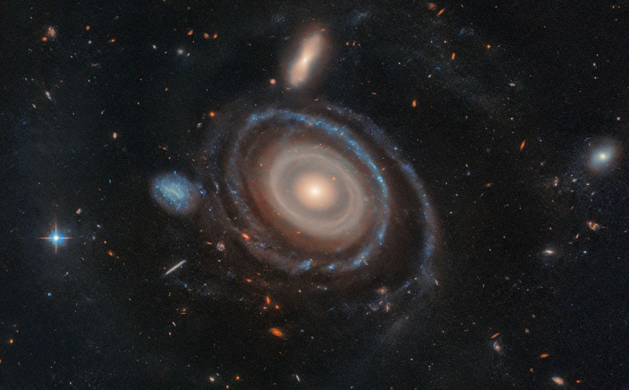 A circular galaxy surrounded by many rings, with several smaller galaxies visible nearby