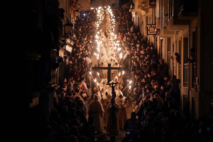 A procession of people wearing white robes and carrying torches walks down a narrow street, surrounded by lookers on either side.