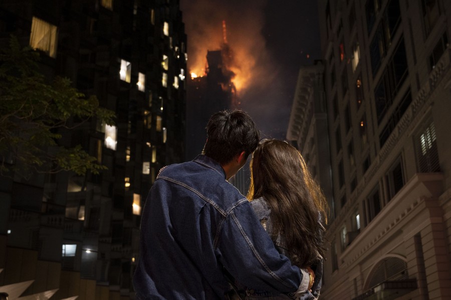 Two people embrace, looking up toward a tall burning building.