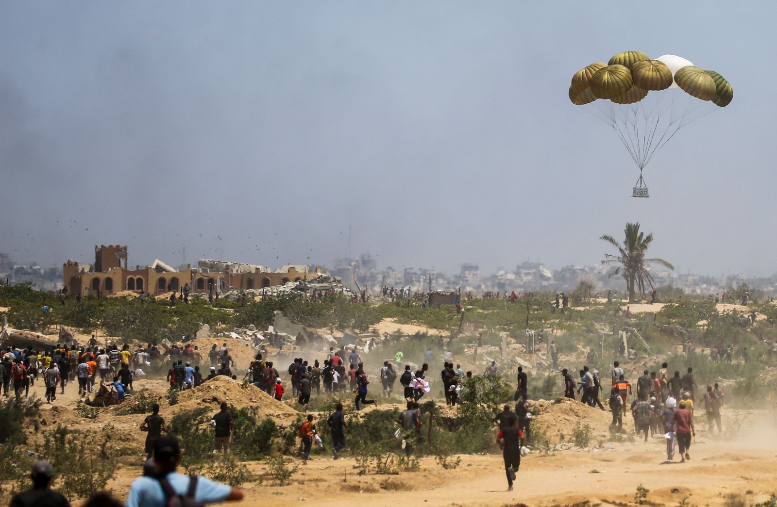 Dozens of Palestinians run across dirt and scrub toward a crate of aid that is parachuting to the ground.