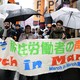 Migrant workers march together on the streets of Tokyo holding banners and umbrellas.