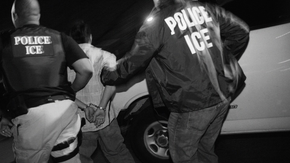 Black-and-white photograph of two ICE officers escorting a handcuffed detainee beside a white van