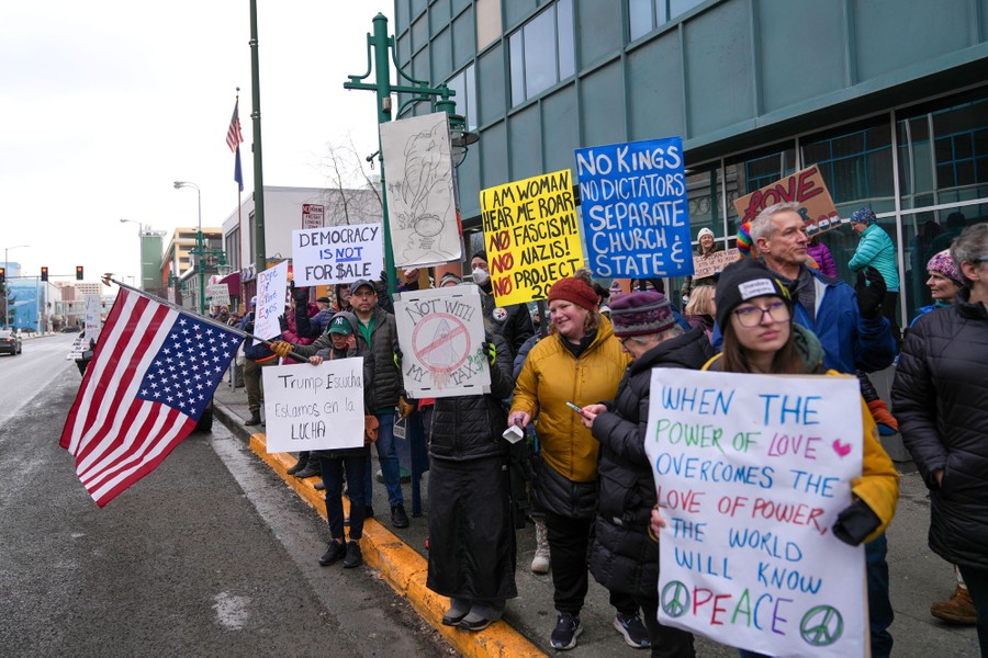 A group of protesters hold signs and flags on a sidewalk, including one that reads 