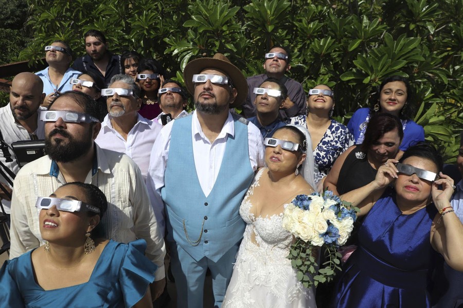 A wedding party stands outside and looks up toward the eclipse, all wearing protective glasses.