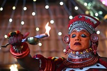 A performer wears an elaborate costume, headgear, and orange makeup.
