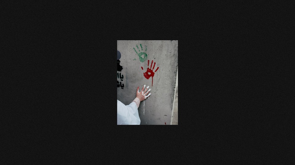 Photograph of a one green and one red handprint on a concrete wall above a bystander with a white-painted hand leaving a third imprint. The photo is surrounded by a thick black border.