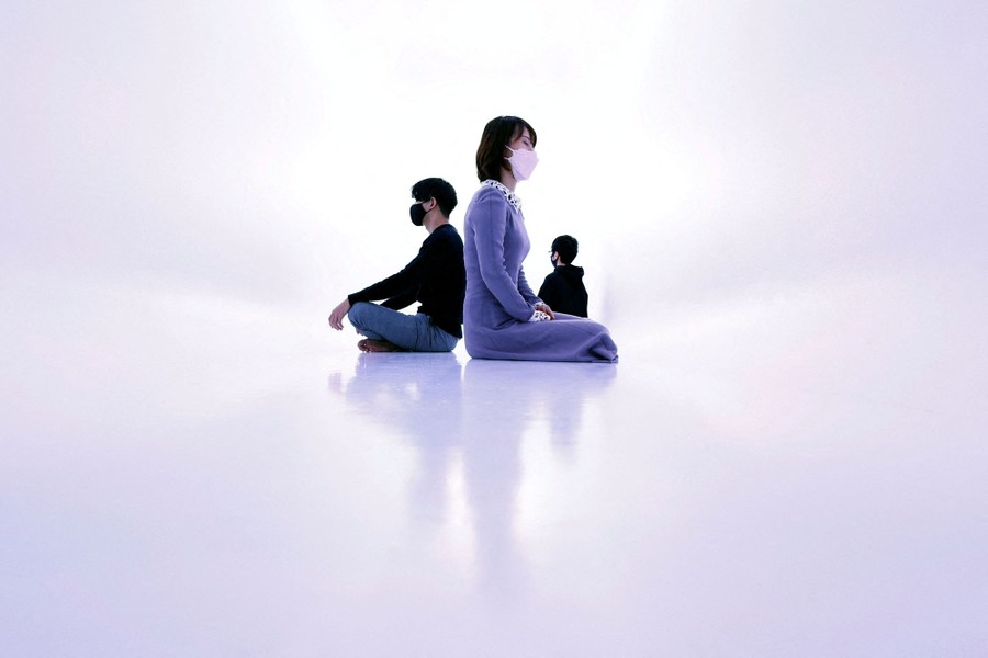Three people are seen sitting cross-legged on the floor of a completely white room.