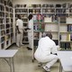 Men dressed in all white sit in a fluorescent room with bookshelves 