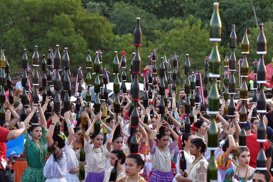 Dozens of people wearing traditional clothing dance while balancing multiple glass bottles on top of their heads.