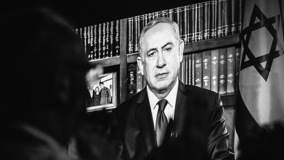 Black-and-white photo of an onlooker watching a speech by Benjamin Netanyahu on a screen.