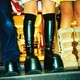Three people are wearing boots while standing on an escalator. The image is taken from the thigh down, with a mall in the background