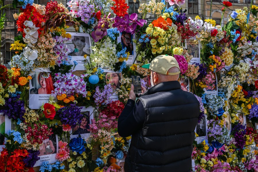 A man stands in front of a temporary fence that is covered with floral arrangements and photographs of people.