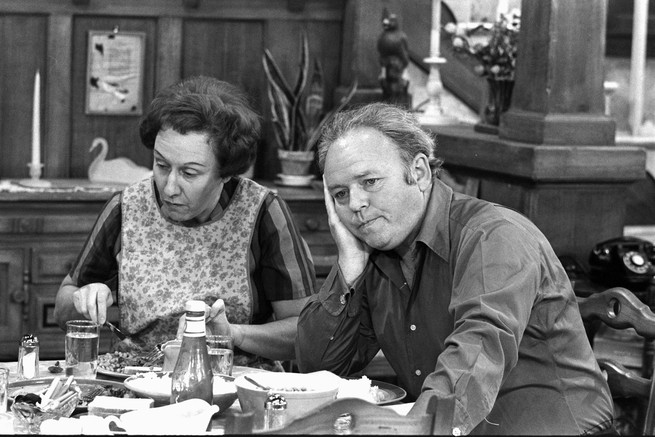 A black-and-white photo shows two of All in the Family's stars, Jean Stapleton and Carroll O'Connor, sitting at a table. 