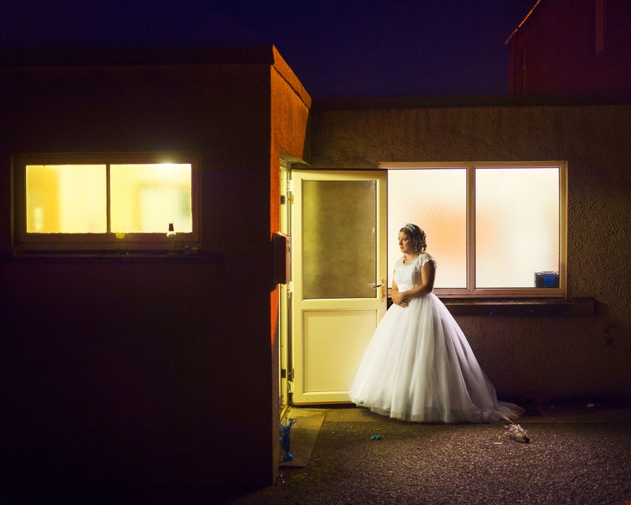 A woman in a long white gown stands outside at night, near an open door.