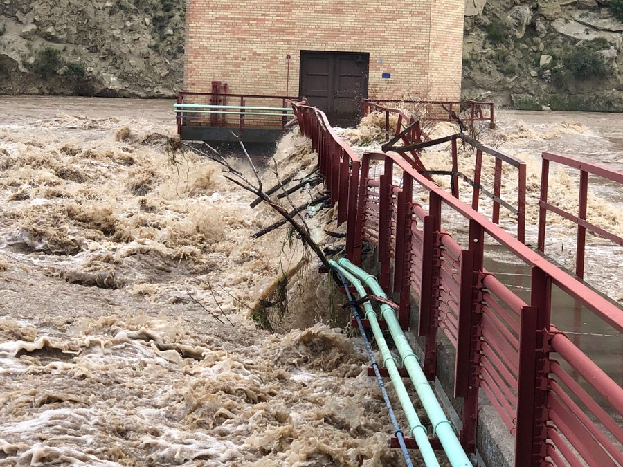 Rushing floodwater overtops a walkway to a brick structure in a river.