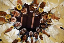 Fourteen athletes, gathered in a circle, all holding gold medals, cheering and smiling down toward the camera.